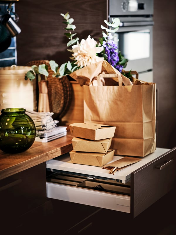 A green KONSTFULL vase on a worktop next to an open UTRUSTA pull-out work surface with a brown paper bag on top.