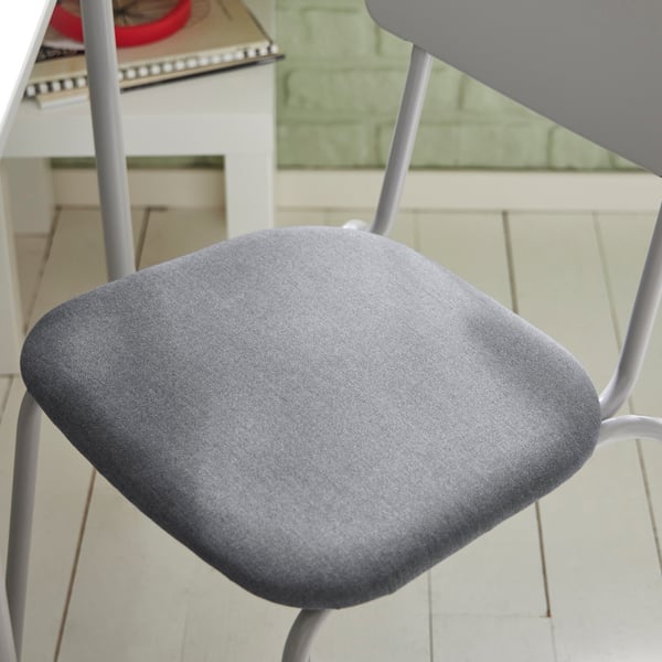 Detail šedé jídelní židle GRÅSALA na bílé dřevěné podlaze, vedle stojí malý bílý stolek se sešitem.