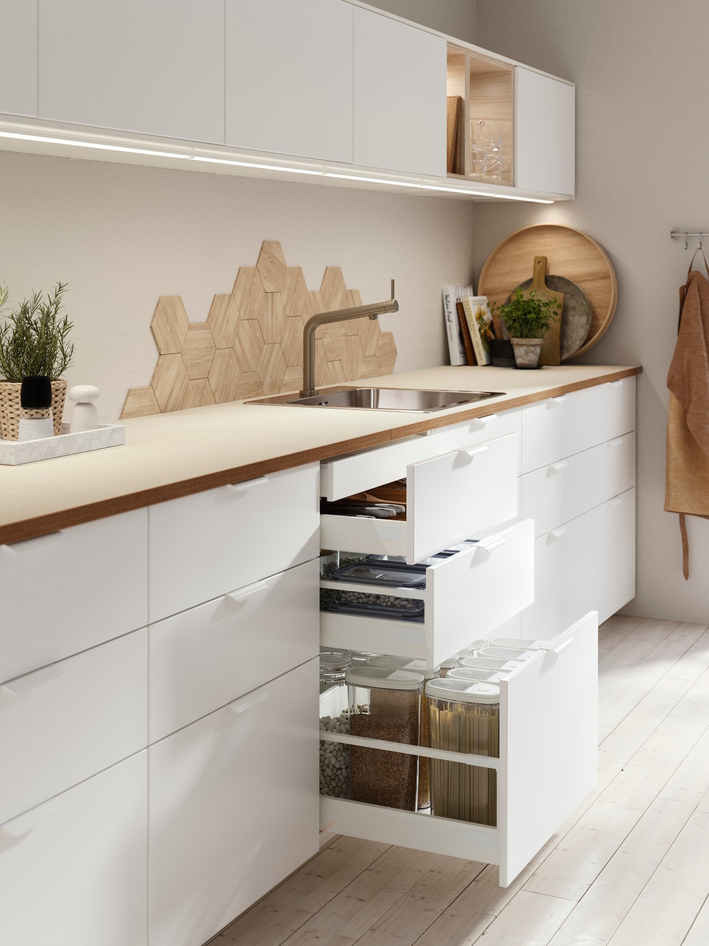 白いキャビネットと木製天板のキッチン。引き出しが開き、シンクと調理器具、壁に飾られたまな板が見える。