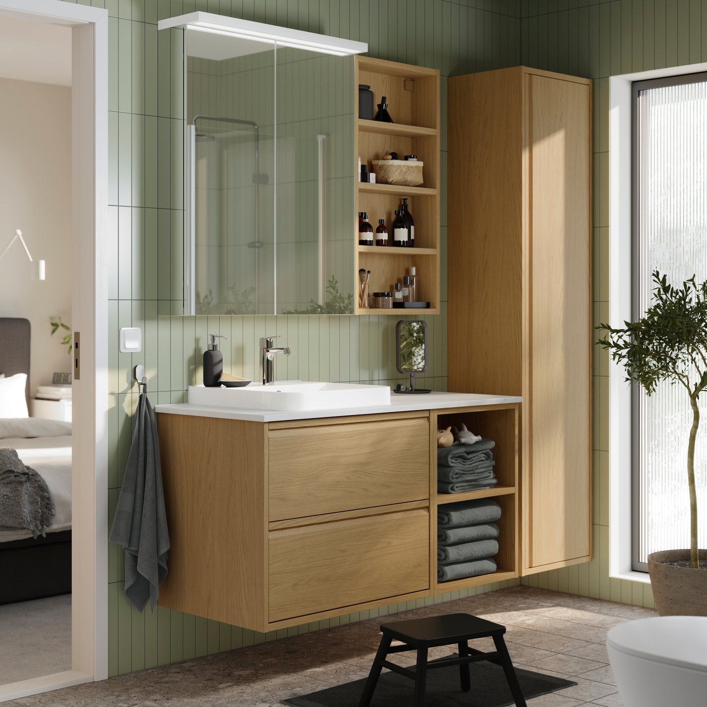 Dans une salle de bains au carrelage vert, il y a un meuble lavabo ÄNGSJÖN effet chêne avec un élément ouvert pour ranger les serviettes grises et une armoire haute.