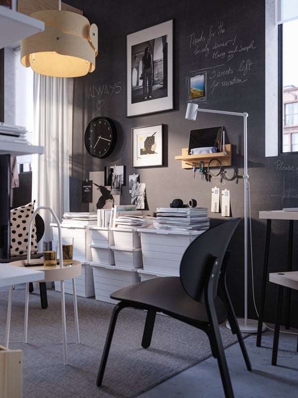 A black FRÖSET easy chair by three stacks of different-sized white SOCKERBIT boxes with lid against a black chalkboard wall.