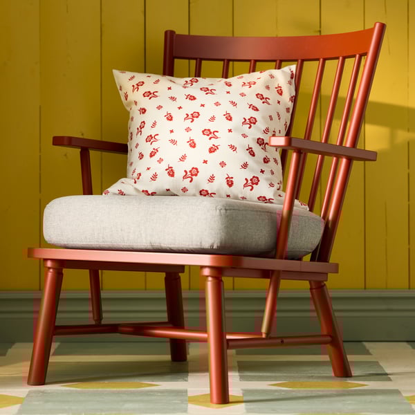 A PERSBOL armchair with a TÅGSÄCKMAL patterned cushion cover sits against a yellow wall, featuring a decorative design.