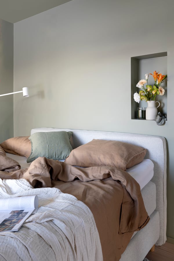 Gemütliche Schlafzimmerecke mit ungemachtem Bett, erdfarbenen Kissen, einer gefalteten Decke und einer Wandnische mit frischen Blumen.