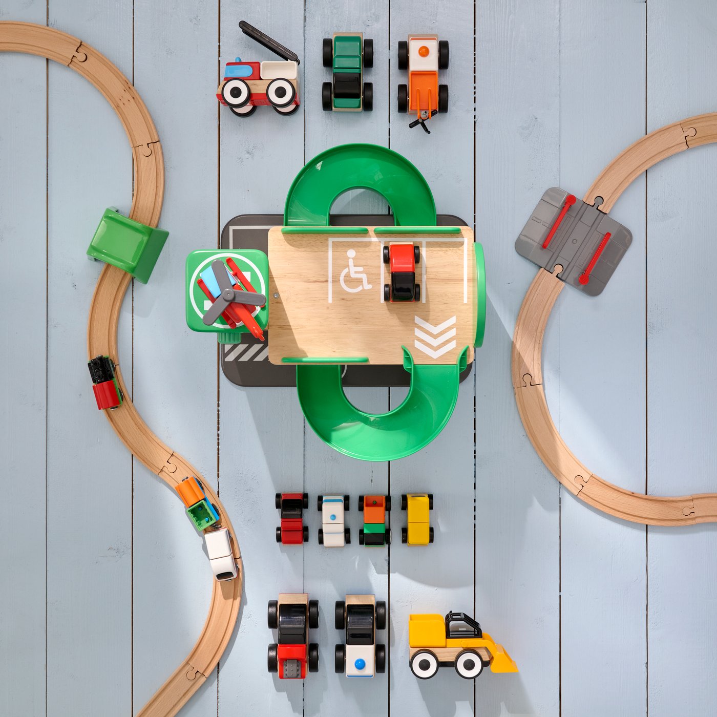 A grey wooden floor of a room shot from above, with LILLABO garage, helicopter, train rail and toy vehicles scattered around.