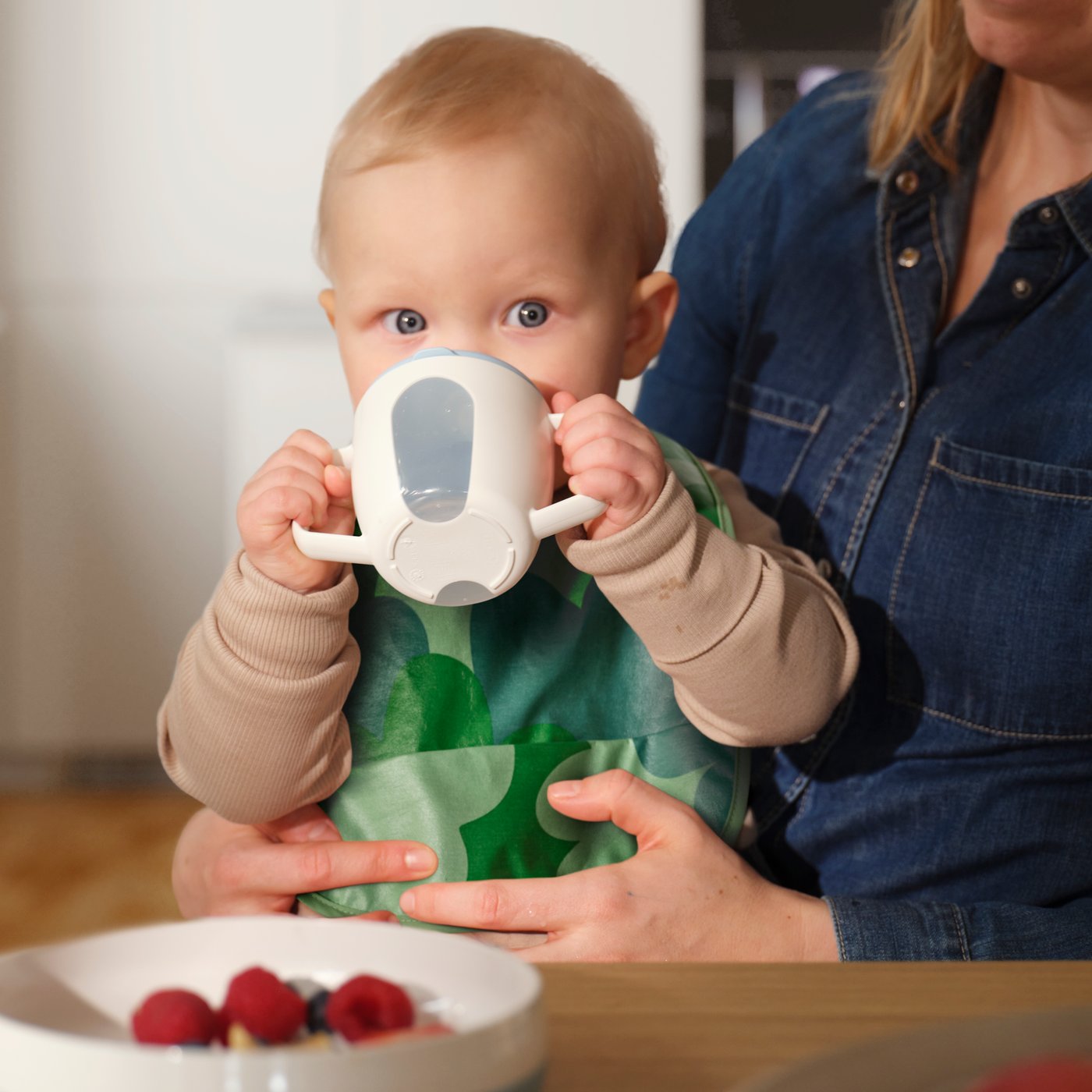 Ett barn sitter i sin förälders knä med en GRÖNFINK haklapp och dricker ur en BÖRJA bägare med en tallrik på matbordet.