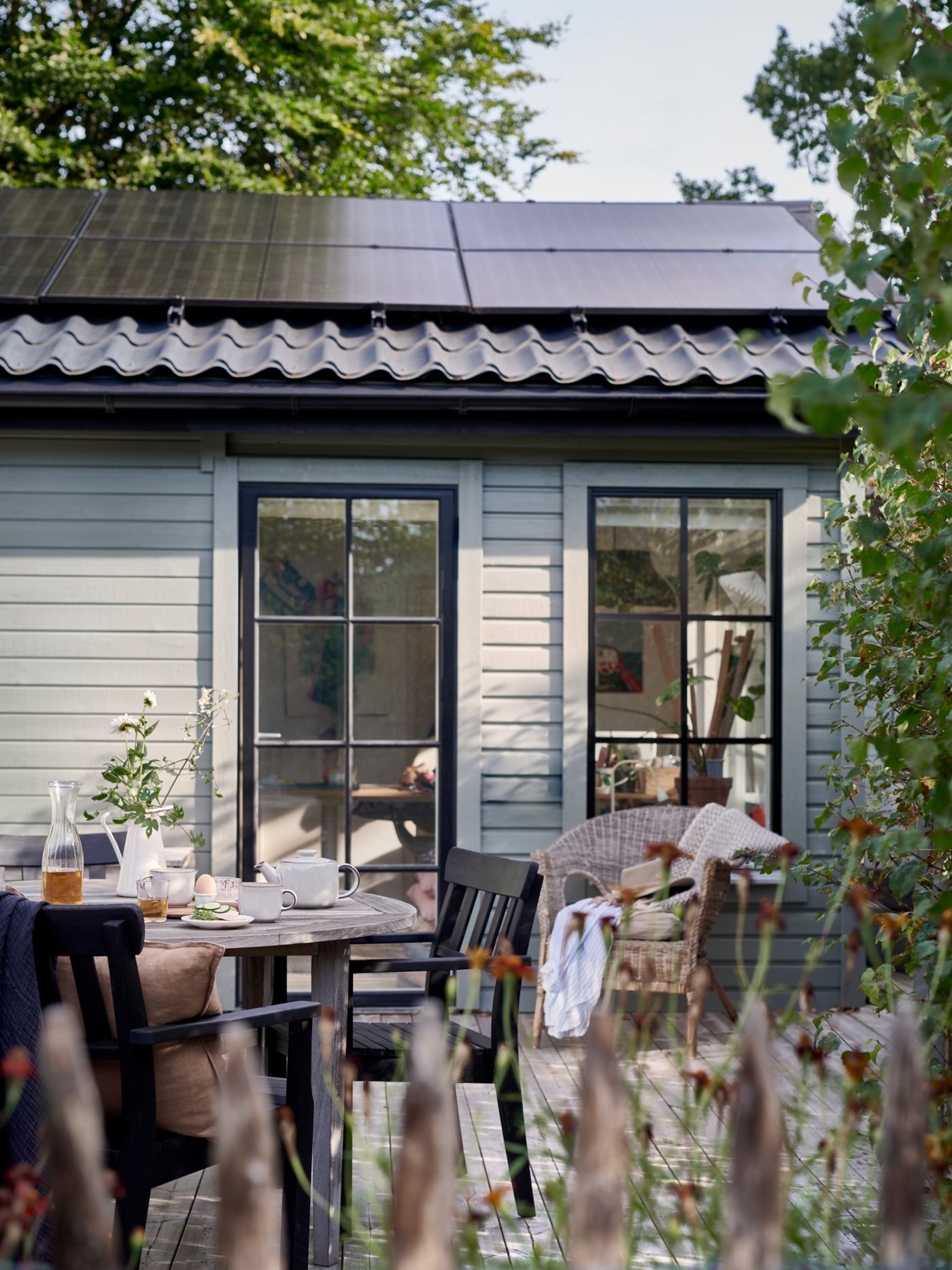 Een schuur of woning met zonnepanelen. We zien de buitenkant van het gebouw: er zijn bomen en een terras.