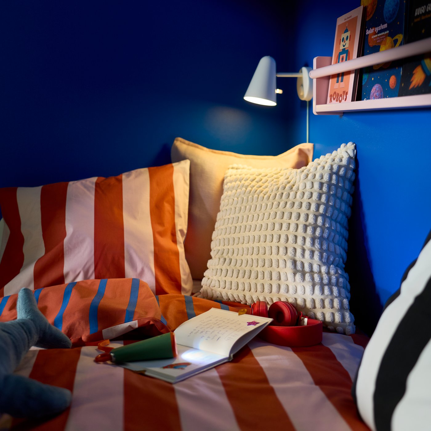 A BARNDRÖM duvet cover and pillowcase set on a children’s bed against a blue wall with a reading lamp and small shelf above.