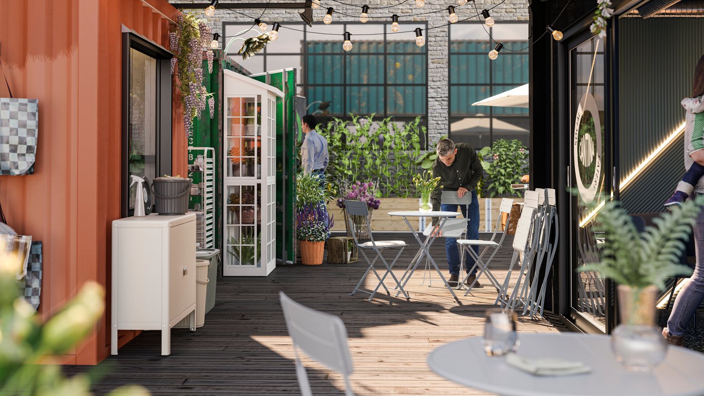 Una cadira SUNDSÖ grisa a la terrassa d'una cafeteria, que està plena de plantes, al costat d'una persona que porta una camisa negra.