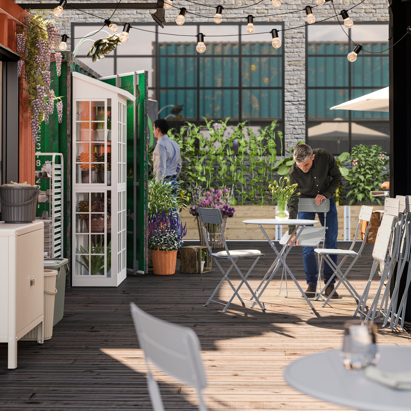 A grey SUNDSÖ chair is being set in an outdoor café area, adorned with plenty of plants, by a person wearing a black shirt.