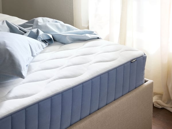 Coin d'un cadre de lit beige avec matelas bleu et blanc et draps bleu clair chiffonnés. Une fenêtre en arrière-plan.