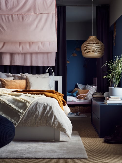 Un lit SONGESAND blanc avec du linge de lit beige et blanc, posé sur un tapis blanc cassé, avec des rideaux roses et une suspension en bambou au-dessus.