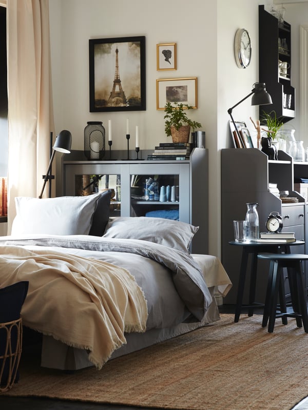 A bed in front of a grey HAUGA glass door cabinet with candlesticks, books and a plant on top. Pictures are on the wall above.