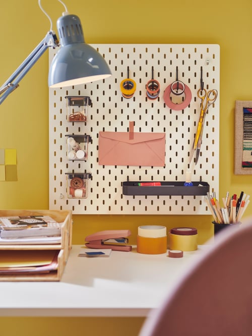 A wall-mounted pegboard with hooks, a container with lid, a shelf, and scissors. A work lamp and a letter tray are on a desk.
