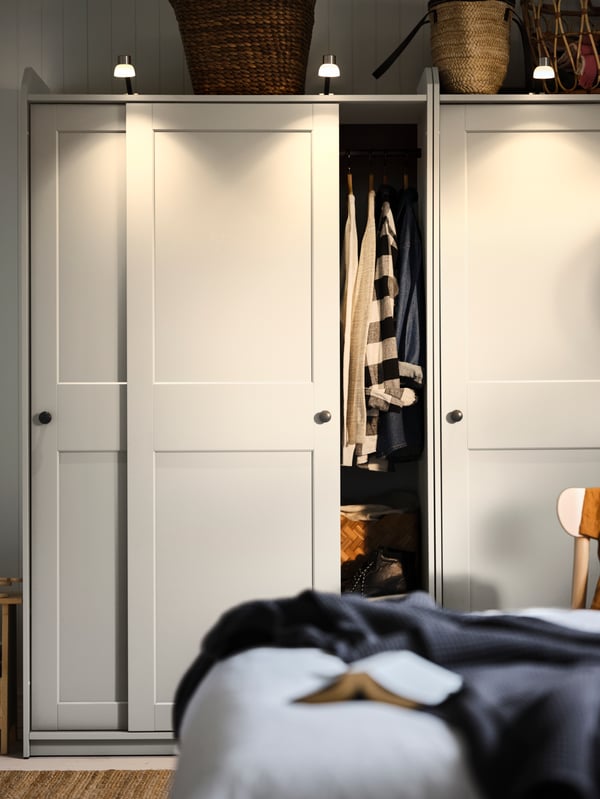 A bedroom where one wall is taken up by white HAUGA sliding-door wardrobes, its front lit up by top-mounted spotlights.