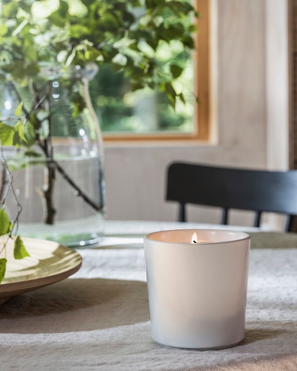 A dining table with an ADLAD scented candle in a ceramic pot and a BEGÄRLIG vase with branches.