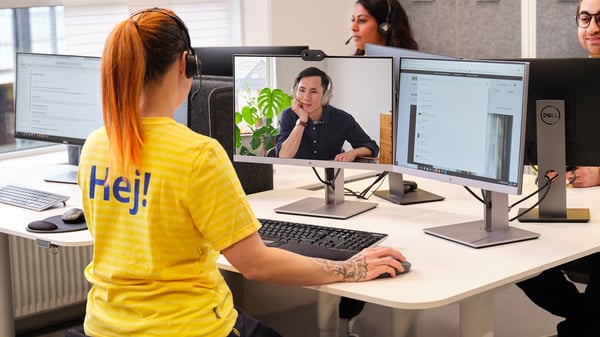 Konsultantka IKEA siedząca przy biurku w żółtej koszulce i prowadząca zdalnie usługę planowania kuchni. Na jednym z ekranów widoczny jest klient korzystający z usługi i rozmawiający z konsultantką.