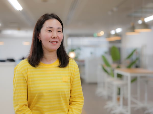 黄色いトップスを着た人物が、観葉植物と白いテーブルが並ぶ明るいオフィスの前に立っている。