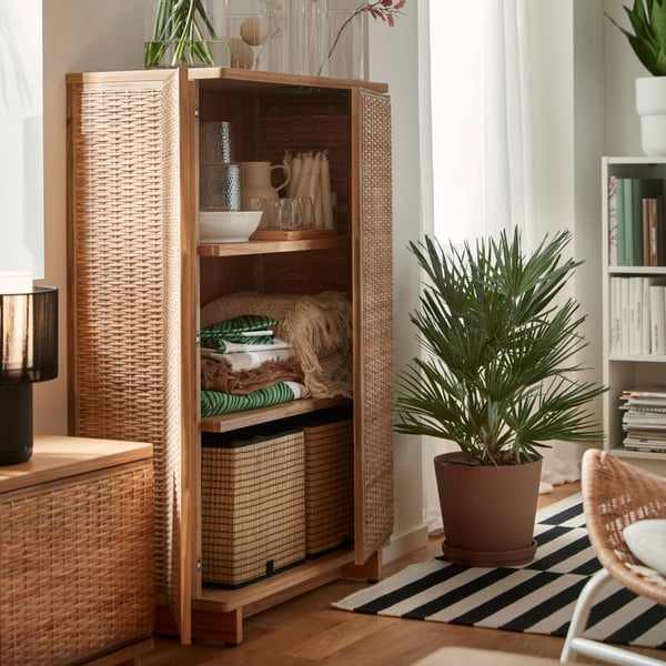 A bright living room with a FRYKSÅS cabinet made of rattan, displaying decorative items and surrounded by plants.