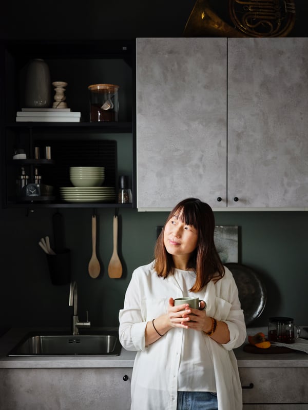A woman drinks a cup of tea in front of ENHET modern concrete kitchen with closed and open storage, in black metal shelving.
