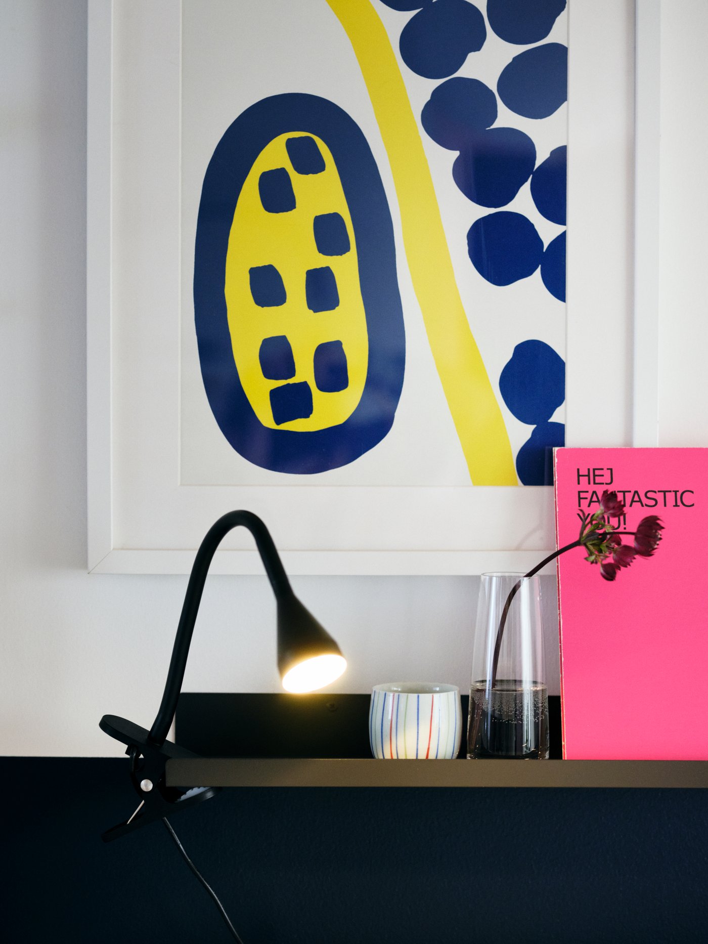 A lit, black clamp spot fastened on a dark shelf, surrounded by wall art, books and decorations.