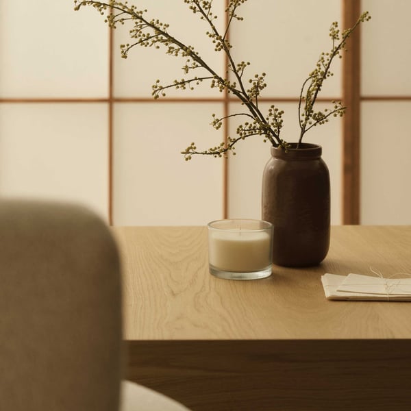 Ett TONSTAD träbord med brun vas med vita blommor, ett vitt ljus i glasbehållare och vikta papper, mot ljusa paneler.