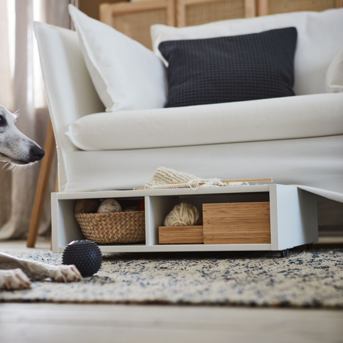 A FREDVANG underbed storage/bedside table is pulled out from under a white sofa next to a dog playing with a ball.
