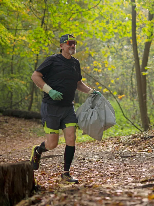 A man is jogging in a forest. He is wearing RIINIG cleaning gloves and carries a trash bag.