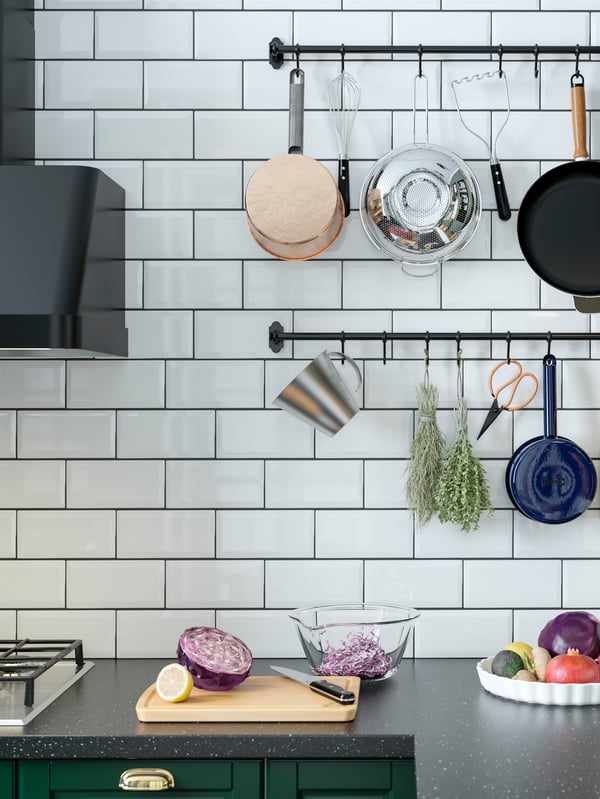 Dua rel berwarna hitam pada dinding dapur berjubin putih di atas permukaan atas kerja. Perkakas dapur dan herba kering tergantung pada rel.