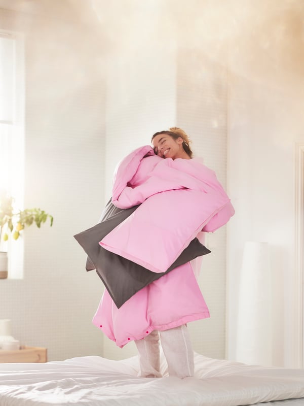Person insvept i rosa PILTANDVINGE påslakan, kramar en grå kudde i ett minimalistiskt sovrum.