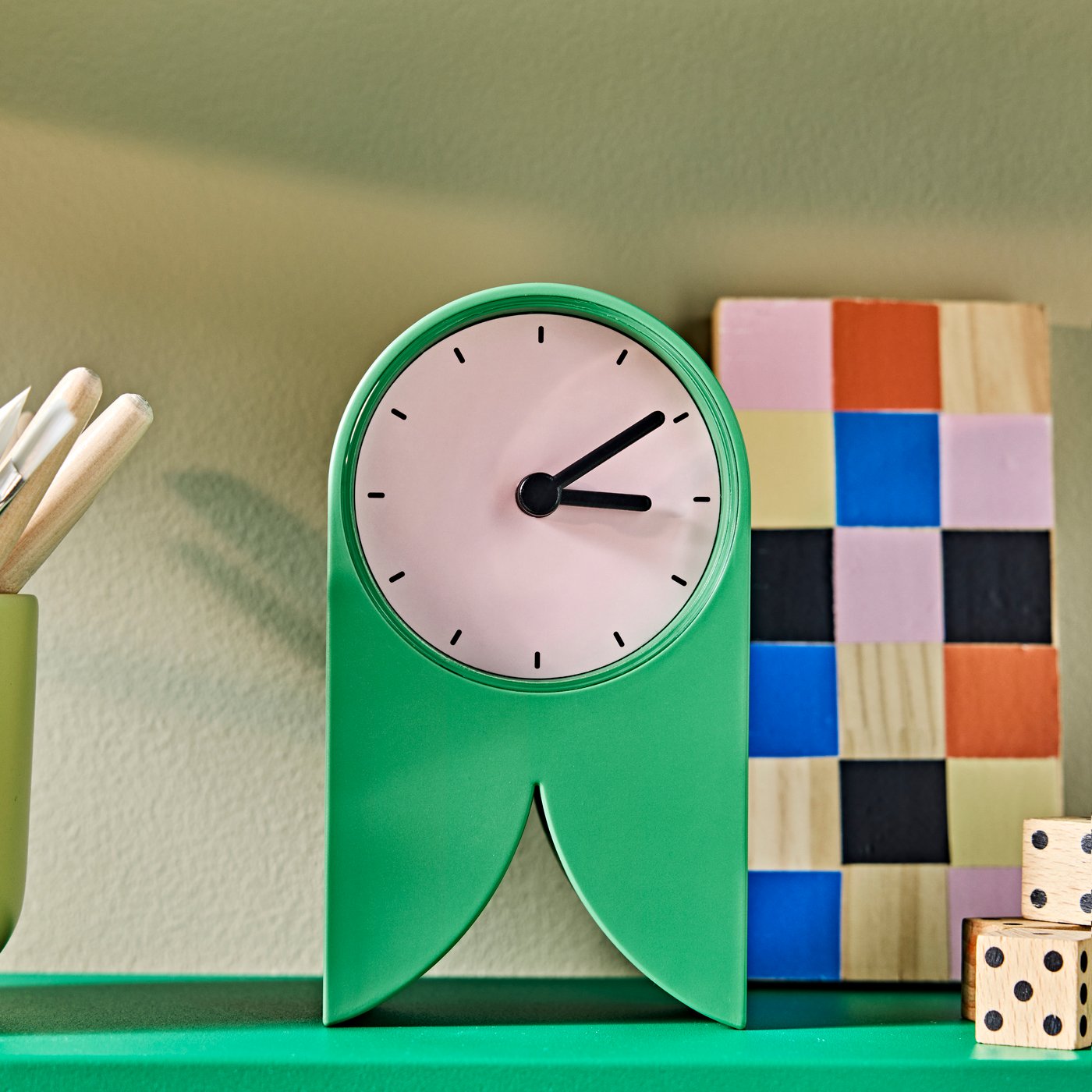 A close-up of a green FLODKRÄFTA table clock with a round face, placed on a shelf beside colourful items.