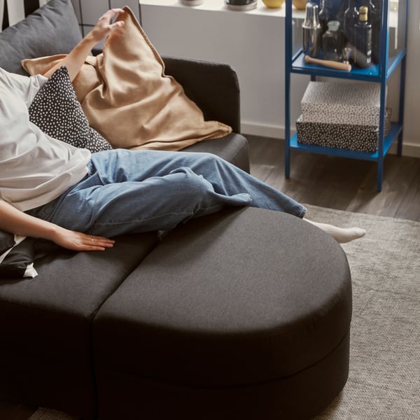 A person wearing jeans is relaxing on a LINDÅKRA two-seater sofa bed in dark grey with a detachable, rounded pouffe.