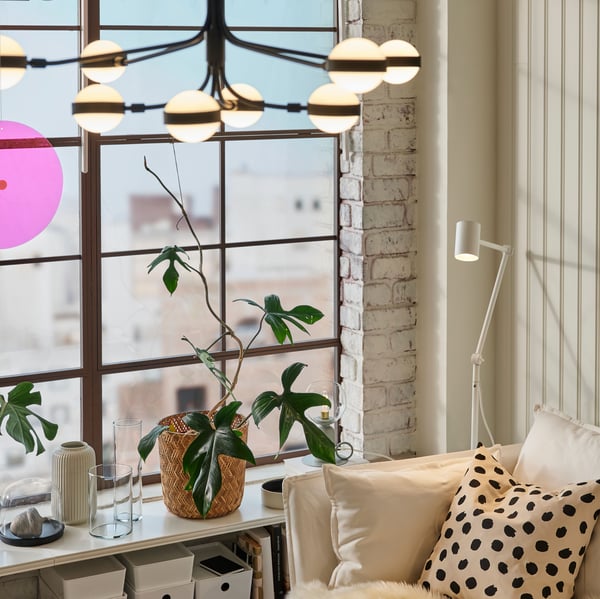 Una lámpara de araña STORSLINGA en blanco y negro en una habitación luminosa con una lámpara de pie NYMÅNE blanca junto a un sofá blanco con cojín.