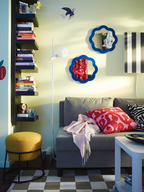 A colourful corner with a black-brown LACK wall shelf with book stacks, an ÄLVDALEN sofa-bed and a yellow EMMISLÖV pouffe.
