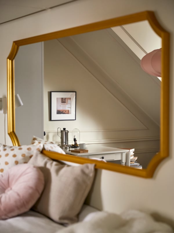 A SVANSELE gold-frame mirror at the head of a bed with cushions in various colours, plus a white table in the reflection.