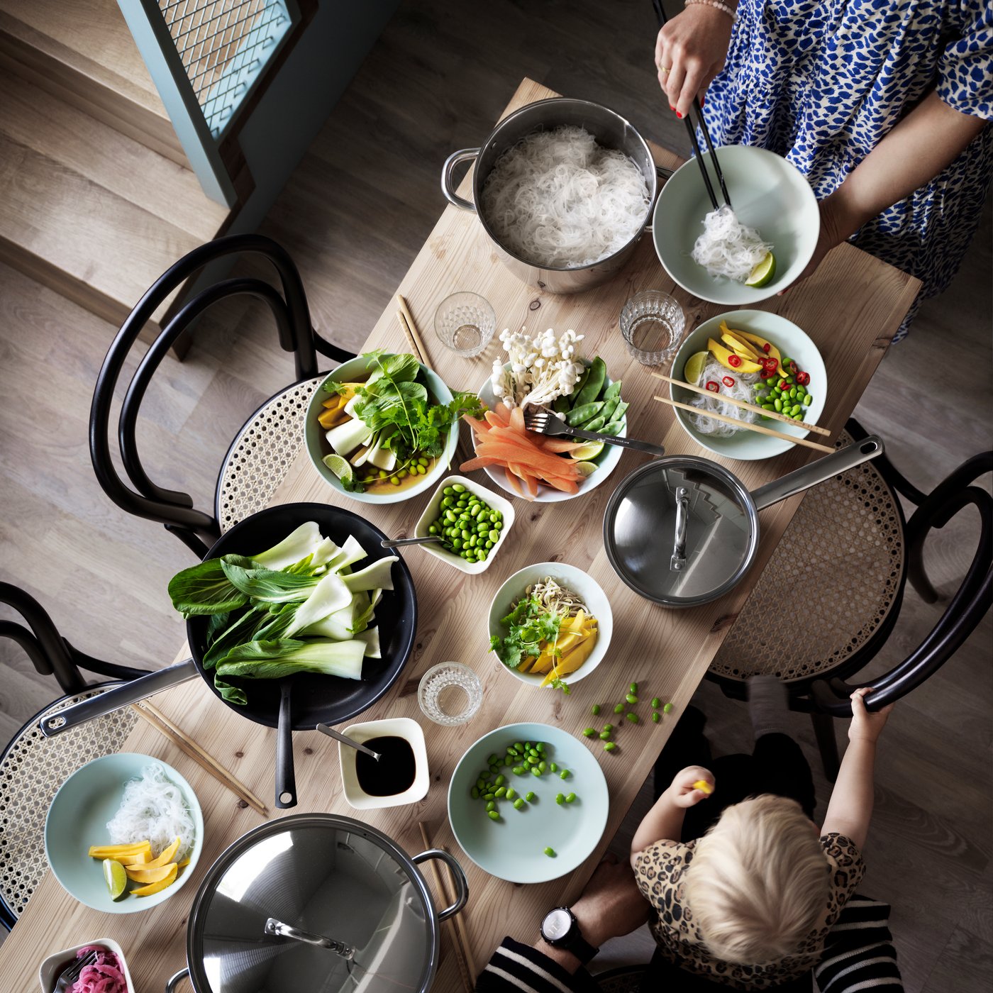 A child sits on an adult’s lap eating finger food at a wooden table with fruit and vegetables in bowls and pans.
