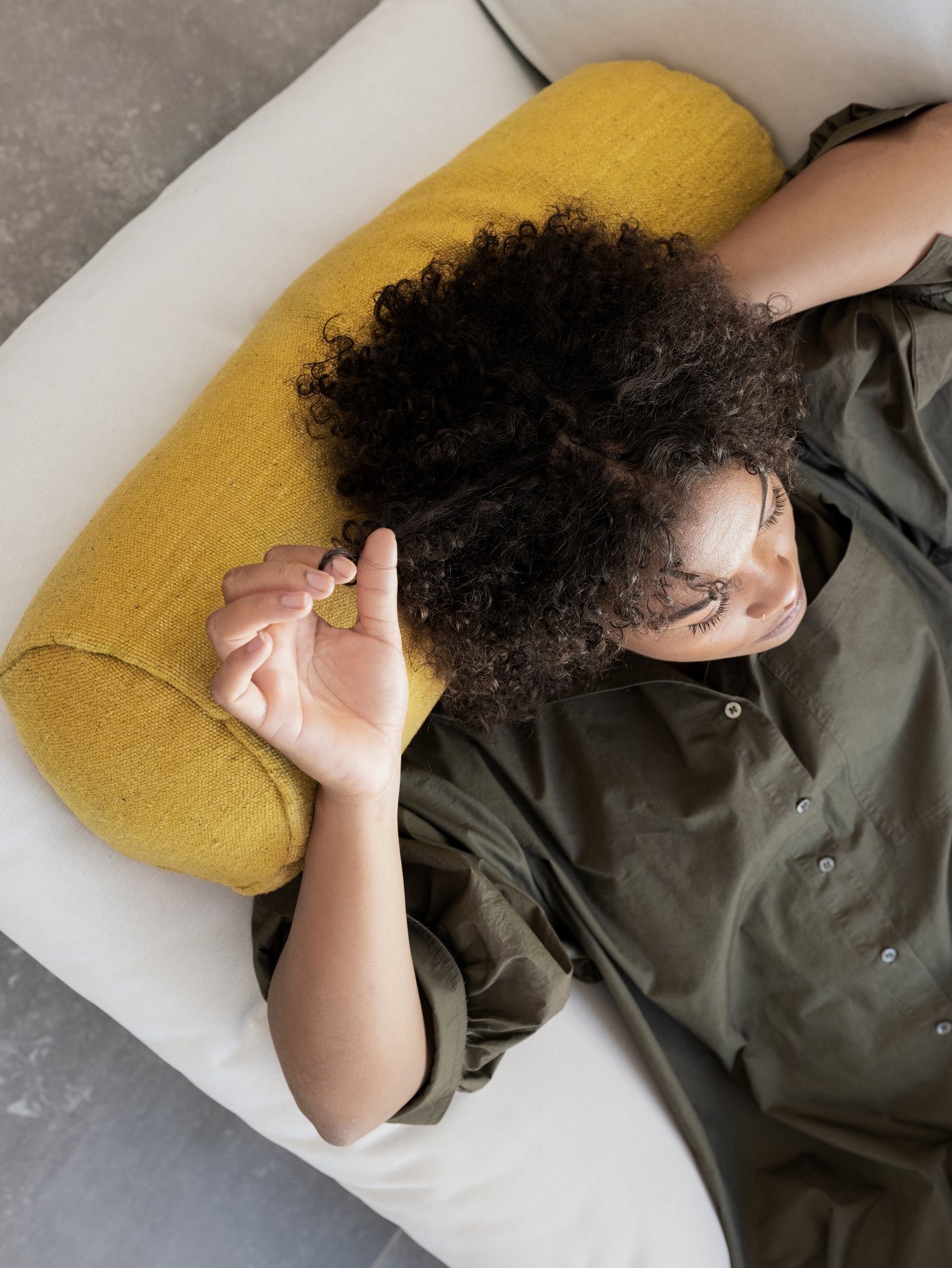 A woman in a brown shirt dress is lying down on a white sofa resting her head on a yellow LOKALT cushion.