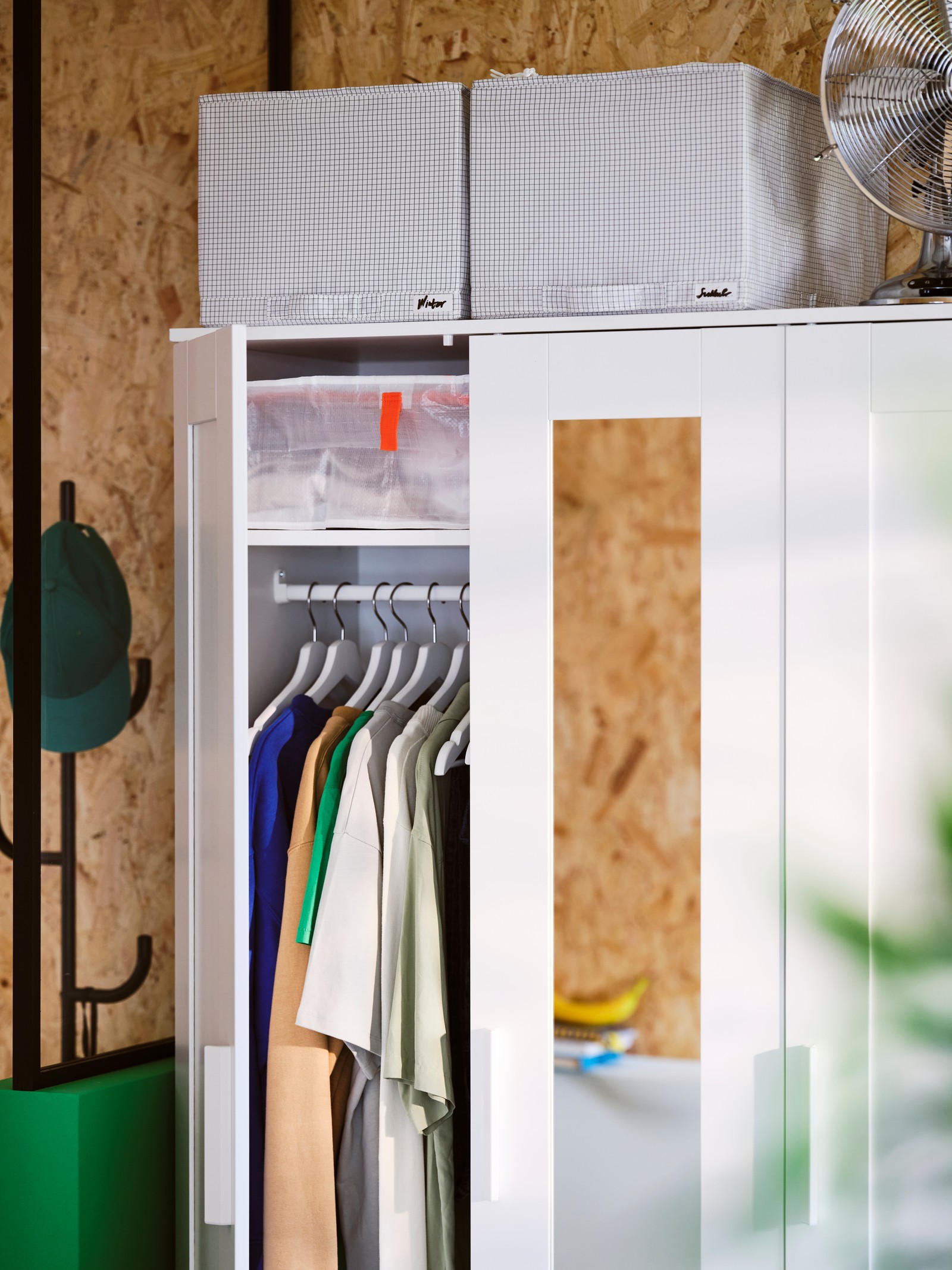 A white BRIMNES wardrobe with 3 doors has one of its doors open showing hanging clothes. Two STUK storage cases are on top.