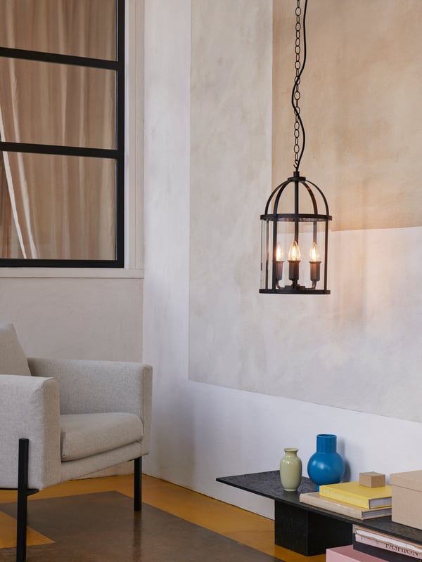 A black, traditional GALJON pendant lamp with three lit decoration bulbs hanging next to an armchair and a coffee table.