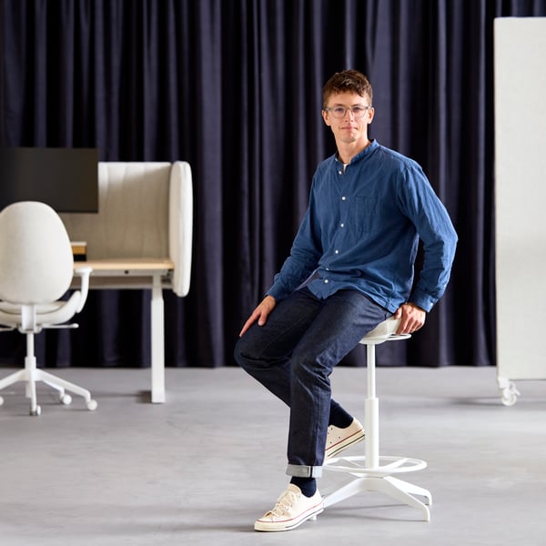 Philip Dilé, product developer, sits on a beige LIDKULLEN active sit/stand support, with a sit/stand desk in the background.