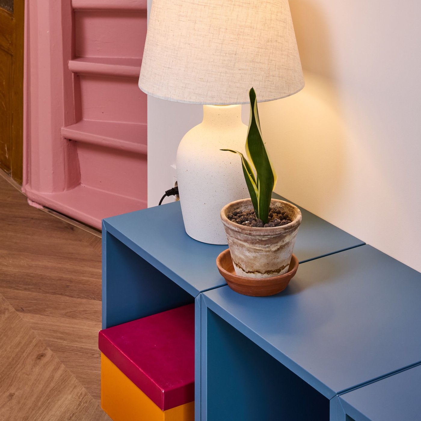 Blauw kastje met witte lamp en kleine plant in pot, kleurrijk ladeblok eronder, houten vloer en roze trap leuning op de achtergrond.
