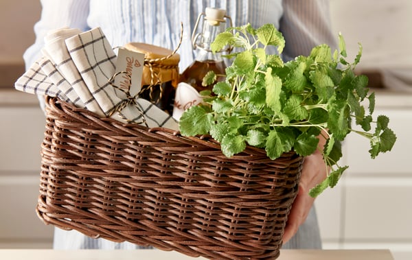 Una donna tiene in mano un cestino GABBIG pieno di strofinacci legati con un nastro, conserve in contenitori di vetro ed erbe aromatiche.