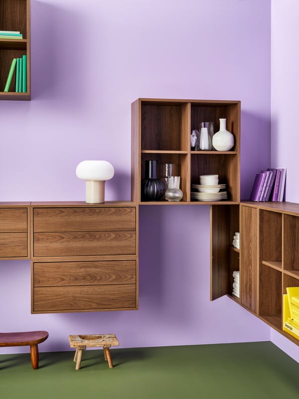 A room with lilac walls featuring different brown EKET cabinets with a walnut effect. There are also books, vases and plates.