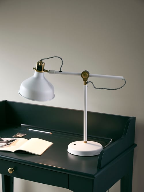 Workspace with white walls, a green desk, a white work lamp, a notebook, black and white photographs and a pencil.