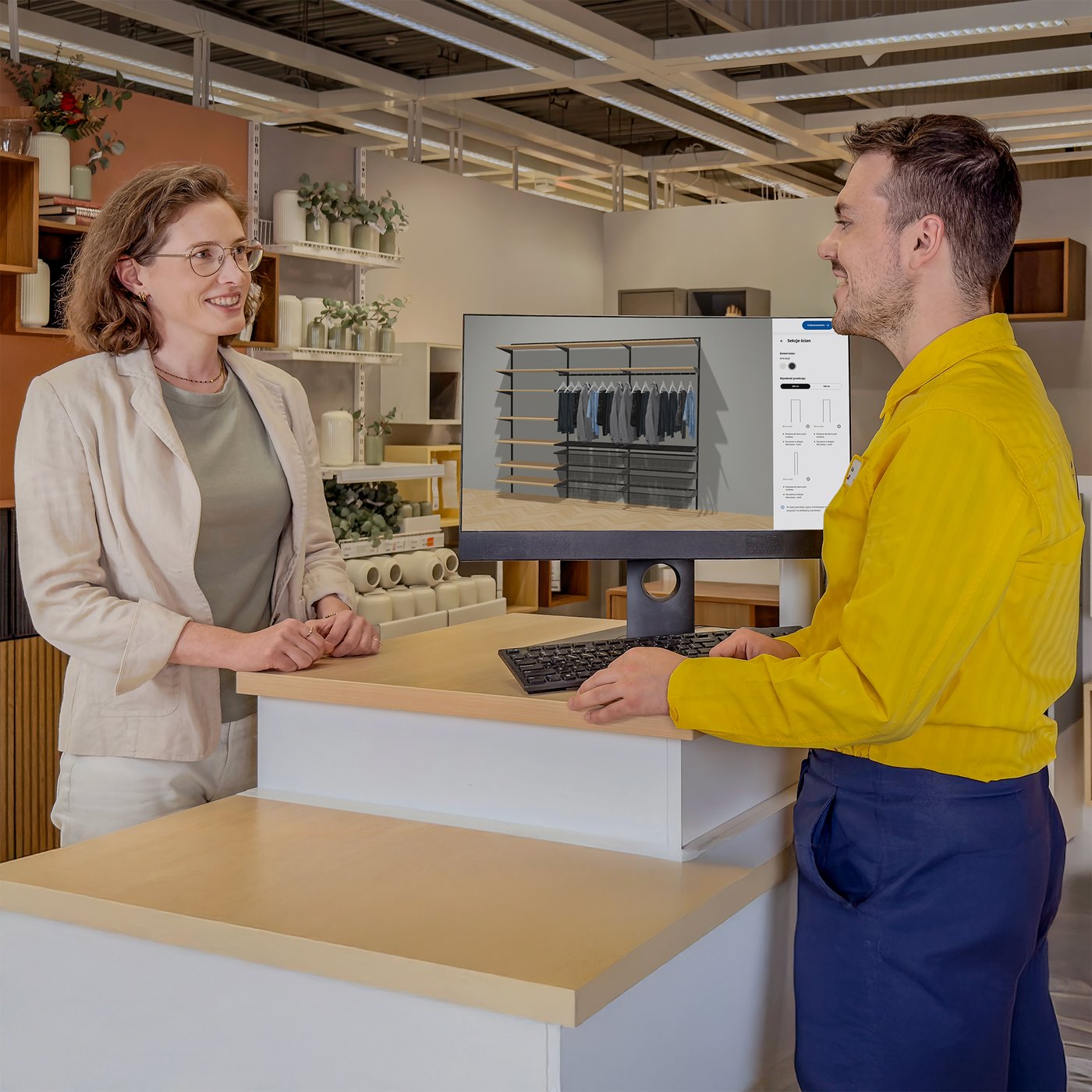 Konsultant IKEA stojący w żółtej koszulce przy stoisku planowania rozwiązań do przechowywania. Przy stoisku stoi również klientka, która omawia z konsultantem projekt rozwiązania na ekranie komputera.