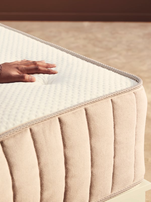 A corner of a VATNESTRÖM pocket sprung mattress with a person’s hand resting on the top surface of the mattress.