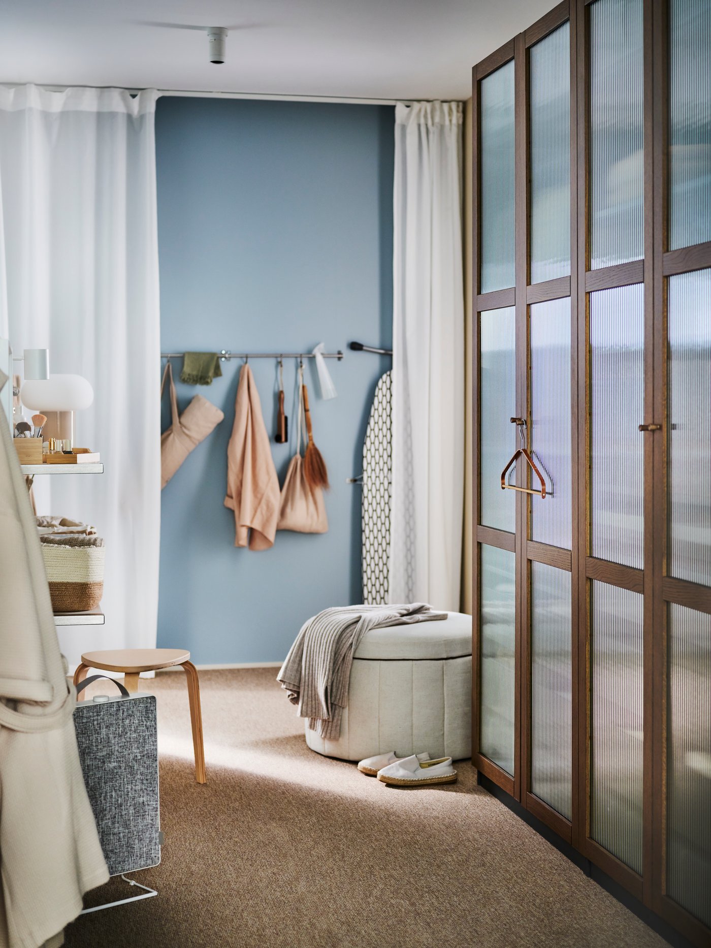 A dark grey brown/stained oak veneer glass PAX/TONSTAD wardrobe combination in a dressing area with a SIDSJÖ pouffe in front.
