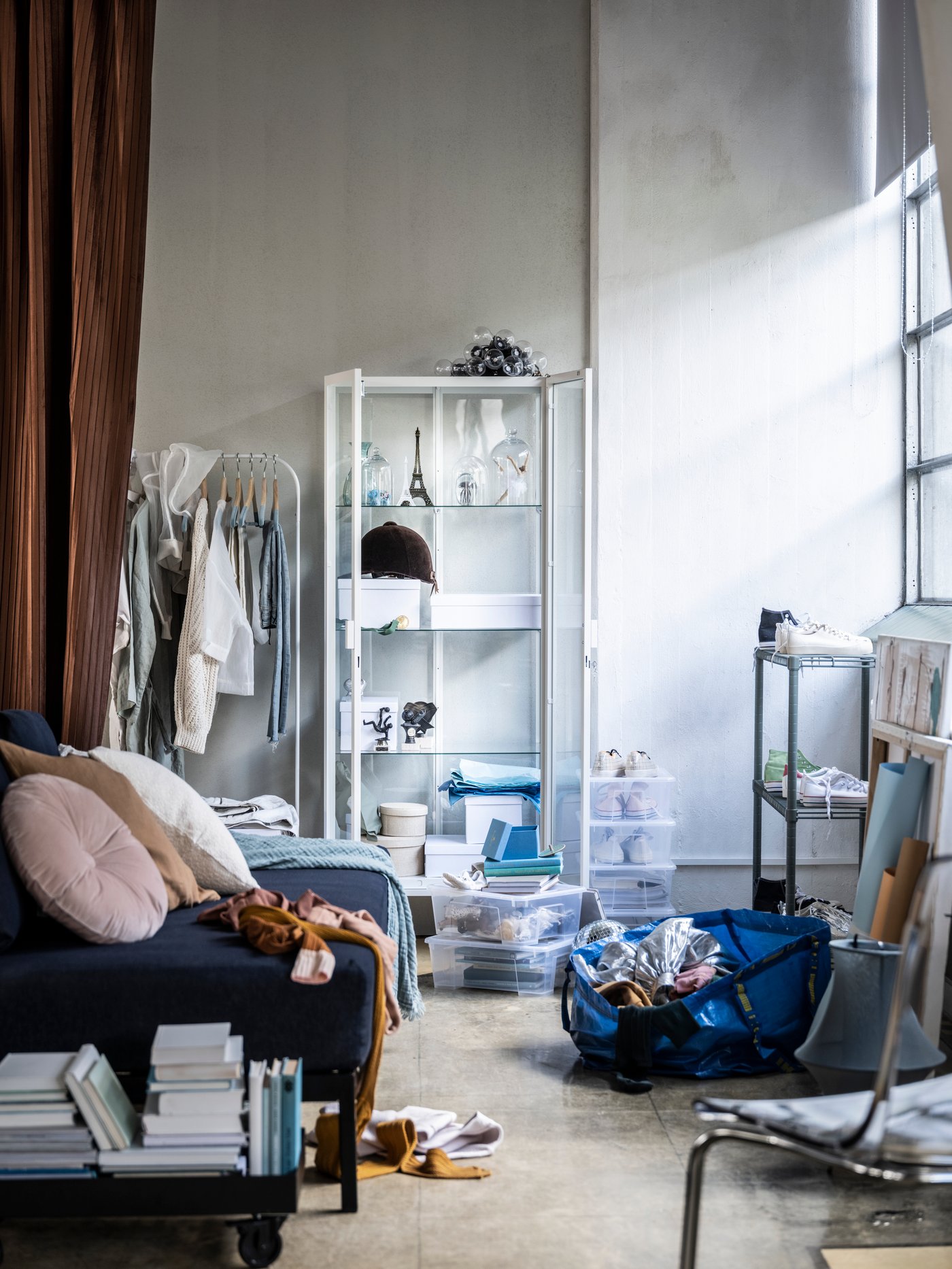 Salón con una vitrina, un perchero, una estantería gris, una bolsa azul, un cojín redondo y ropa.