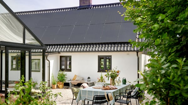 Es ist ein weißes Haus mit einem schwarzen Dach zu sehen, auf dem sich eine Photovoltaikanlage befindet. Das Haus ist von einem üppigen grünen Garten umgeben. Auf einer Terrasse gibt es einen gemütlichen Außenbereich mit einem Tisch mit karierter Tischdecke, Tellern und Speisen und Stühlen.