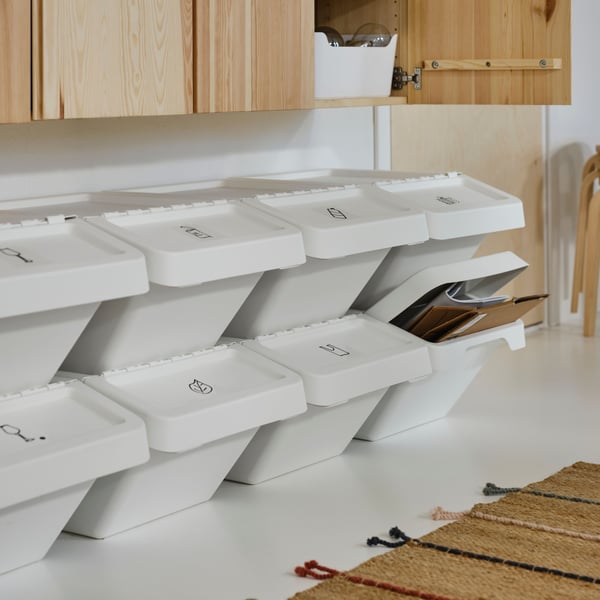 The image displays a tidy storage area with a wooden shelving unit on the left side, containing various items such as terracotta pots, a green watering can, and some cans of paint. In front of the shelving unit are two gray fabric bins with white outlines of a battery and a cardboard box printed on them, suggesting they are for recycling specific types of waste.