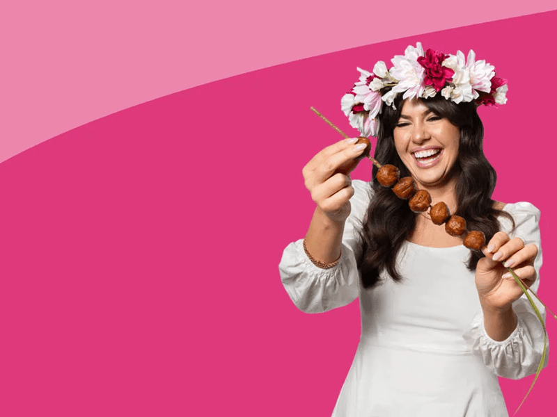 Jeune femme brune souriante habillée de blanc avec une couronne de fleur sur la tête , piquant des boulettes de nourriture sur un pic à brochette sur un fond rose.